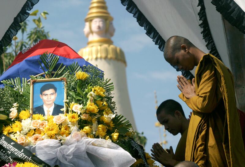A framed photo of Chea Vichea and yellow flowers rest on the union leader's coffin in the left side of the photo. Two buddhist monks bow their heads and clasp their hands in prayer to the right.